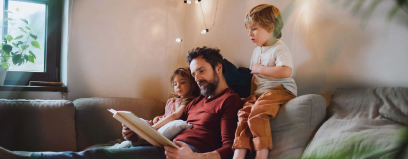 Um pai lendo para seus dois filhos pequenos no sofá, um gesto simples que estimula o gosto pela leitura, fortalece laços familiares e contribui para o desenvolvimento infantil.