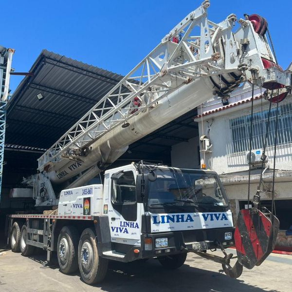 Guindaste telescópico e caminhão munck operando em canteiro de obra industrial com céu azul e equipe técnica no local 