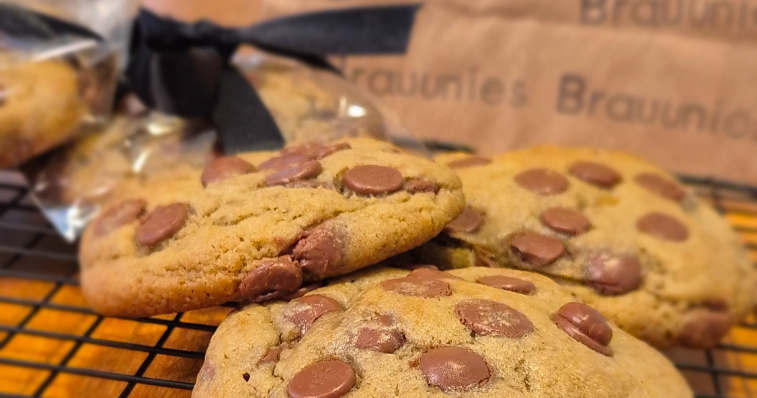 Pote transparente aberto com cookies artesanais de chocolate empilhados sobre mesa de madeira rústica, ao fundo embalagem personalizada e flores secas 