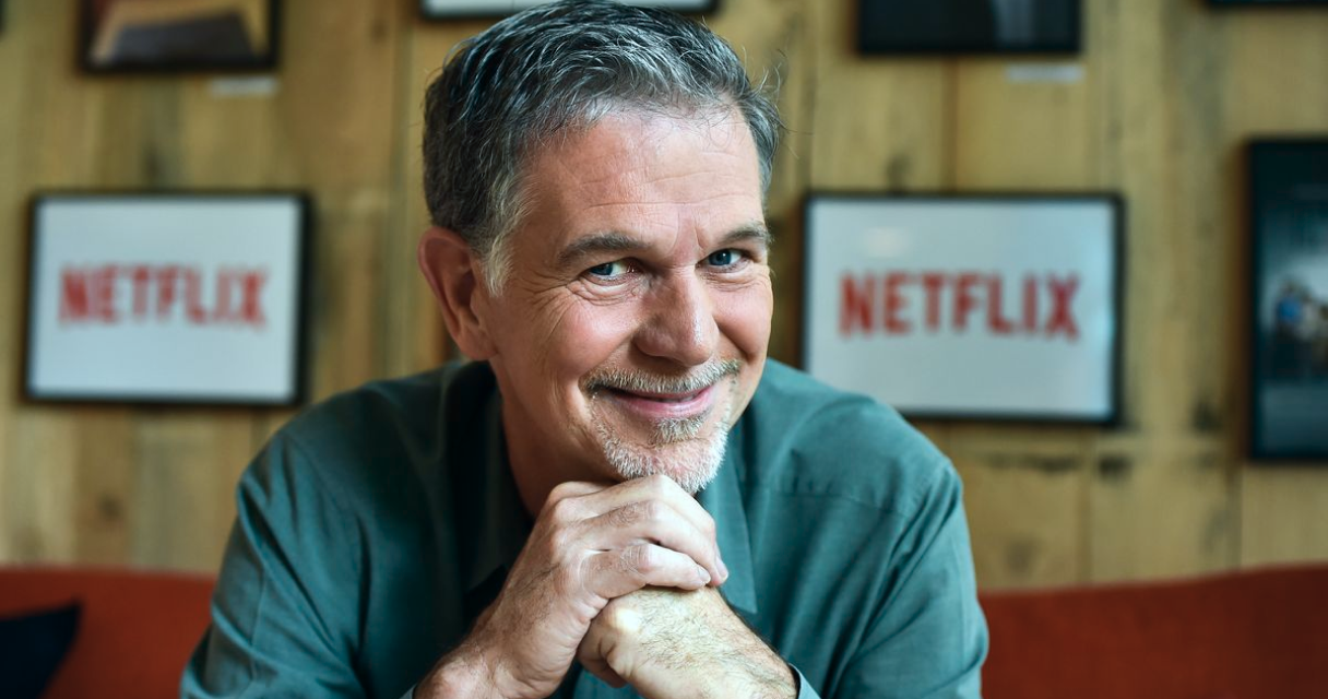 Homem sorrindo em ambiente decorado com quadros da Netflix ao fundo, apoiando as mãos sob o queixo enquanto olha para a câmera.
