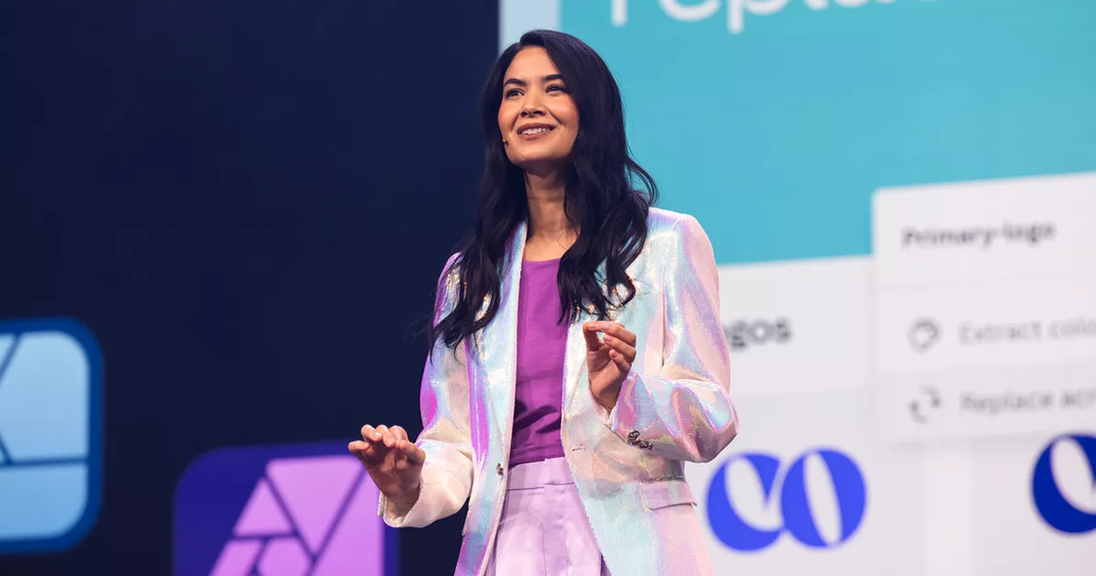 Mulher apresentando em palco, sorrindo, com blazer iridescente e fundo com gráficos coloridos de tecnologia e design.