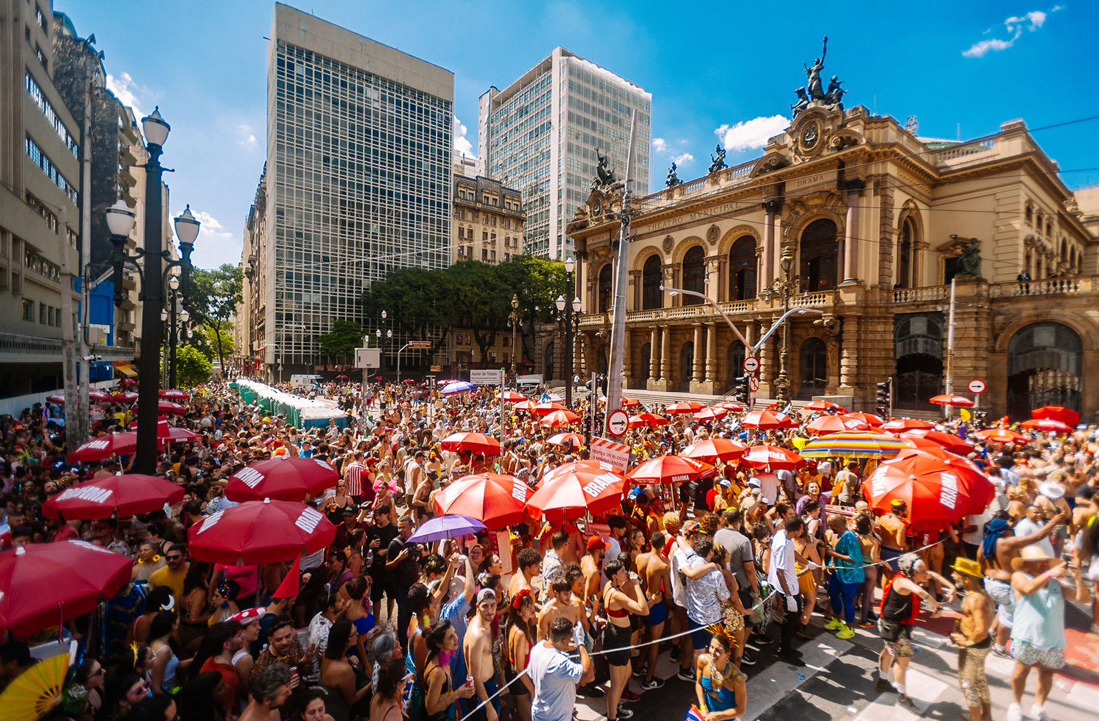 Bloco de rua lotado no Carnaval de São Paulo com foliões fantasiados e prédios ao fundo 