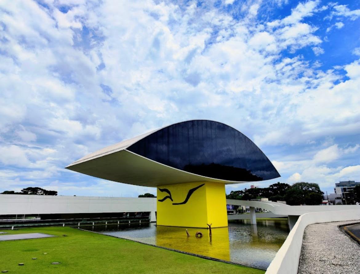 Sedã executivo passa por avenida arborizada com Museu Oscar Niemeyer ao fundo em Curitiba 