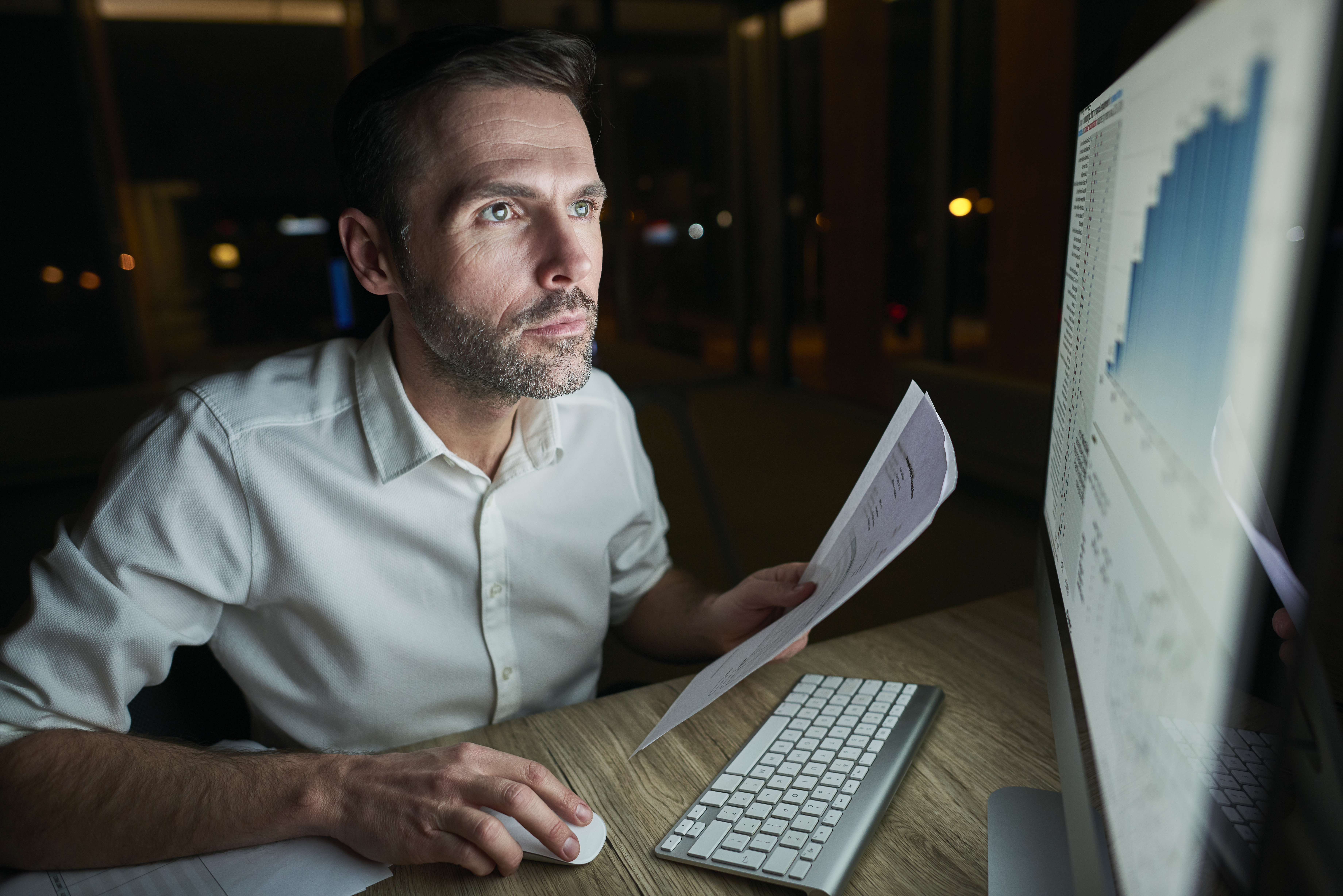Homem olhando indicadores de gestão de dados em seu computador, enquanto segura papéis na mão e, na outra, move o mouse que está sobre uma mesa de madeira